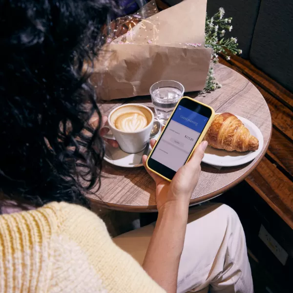 A person sits at a café table with a croissant, cappuccino, and water, holding a smartphone showing a banking app.