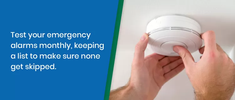 A person tests a smoke detector on the ceiling. Text beside reads: Test your emergency alarms monthly, keeping a list to make sure none get skipped.