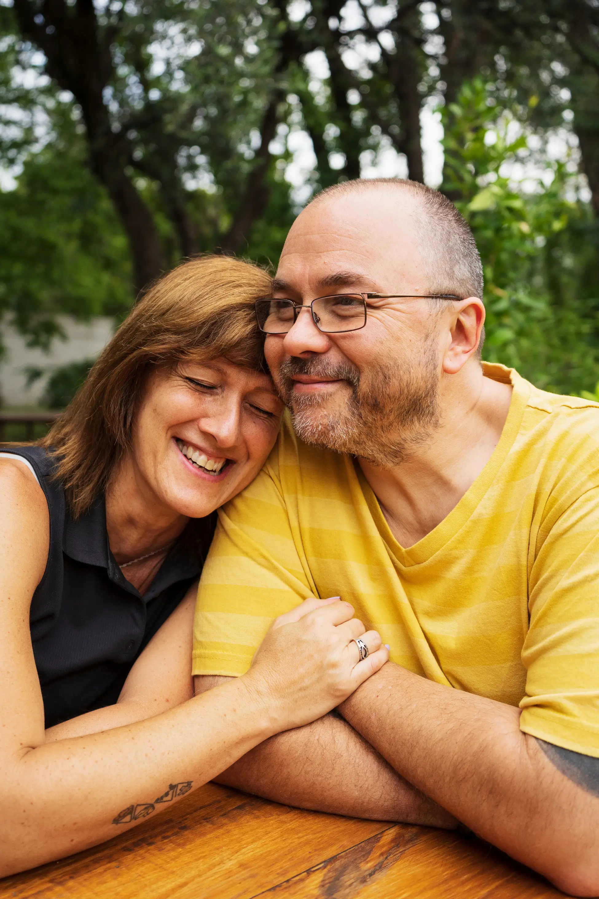 Smiling couple sits close together at a wooden table outdoors, with the woman leaning her head on the mans shoulder, both looking happy and relaxed.
