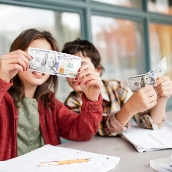 Two children sitting at a table with notebooks, holding and examining U.S. hundred-dollar bills, with a pencil lying on the table.