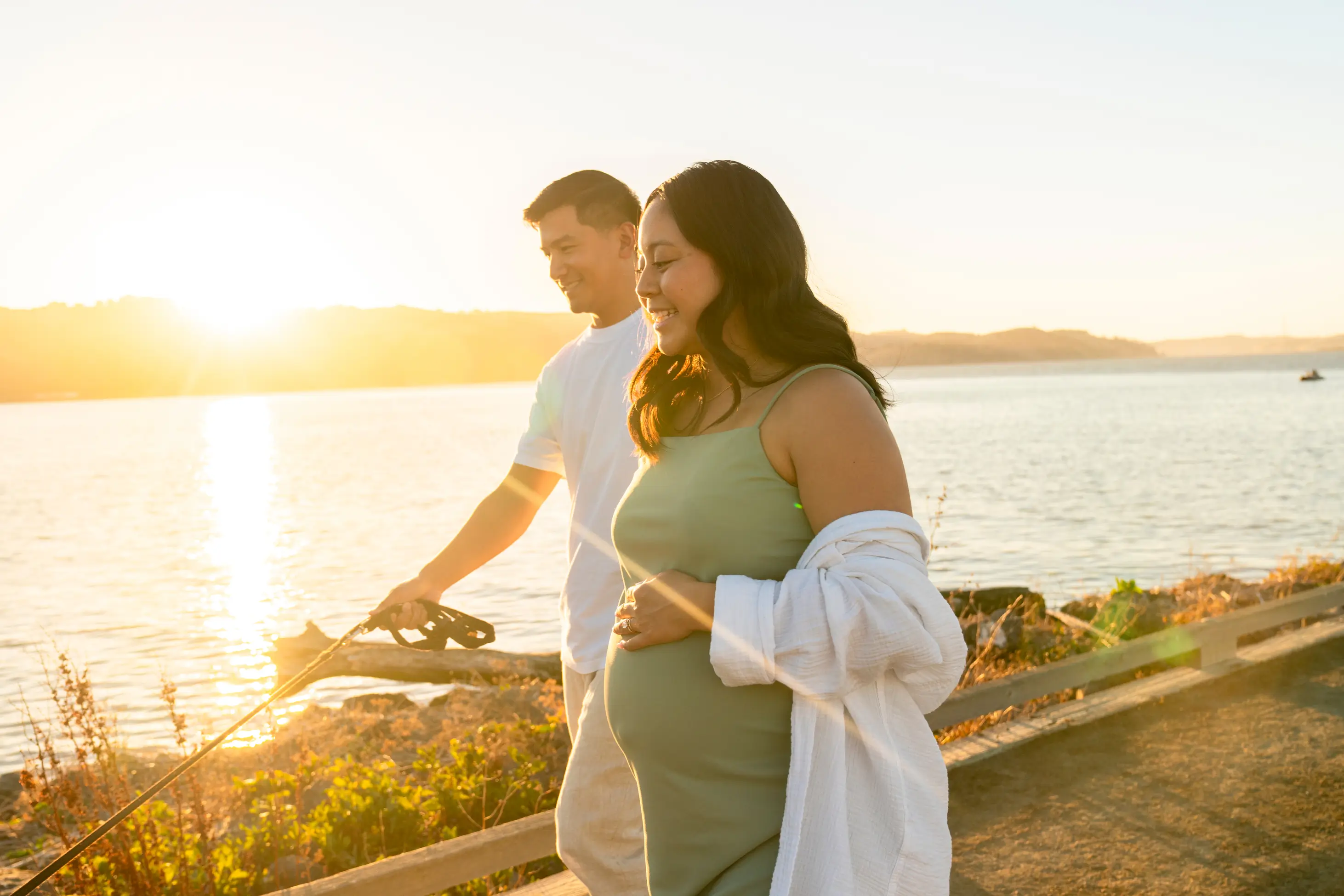 A smiling couple walks a dog near a body of water at sunset. The woman, who appears pregnant, wears a green dress and white cover-up. The man holds the dogs leash.