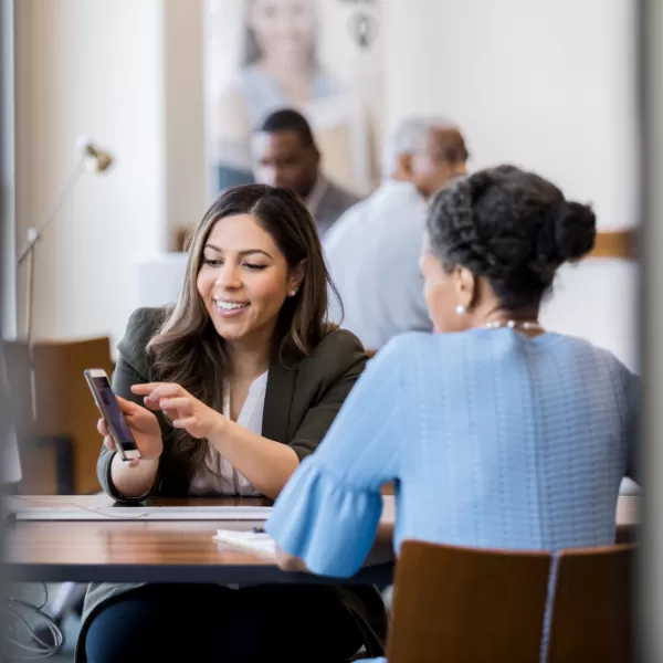 Two women sit at a table in an office, one smiling and showing something on her phone to the other, who listens attentively. Other people are blurred in the background.
