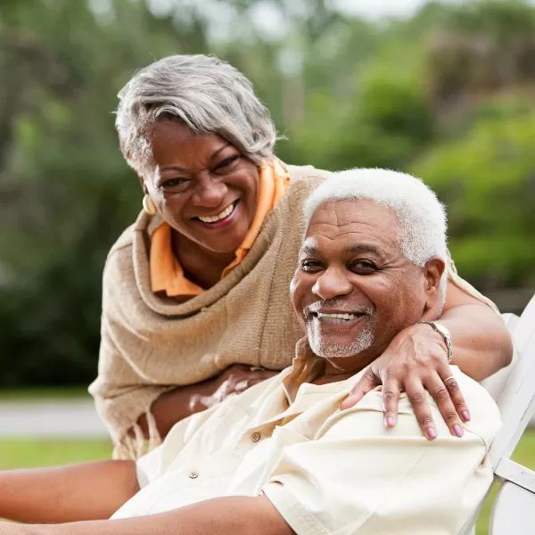 Smiling older couple poses together outdoors, with the woman standing behind the man and wrapping her arm around his shoulder. Both look happy and relaxed.