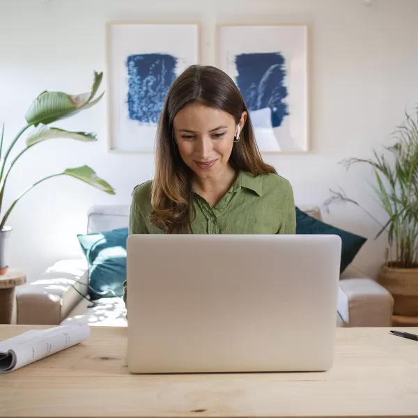 A woman in a green shirt smiles while working on a laptop at a wooden table in a cozy, plant-filled living room.