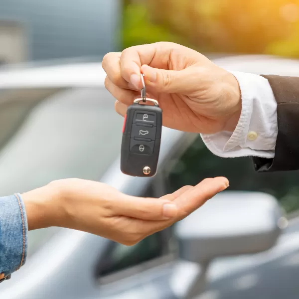 A person hands over a car key to another person with an open palm, with a blurred car in the background.