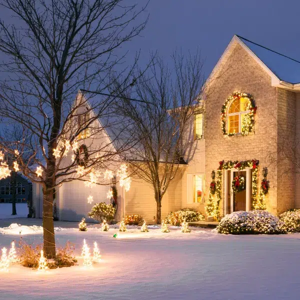 A two-story house decorated with Christmas lights and wreaths, surrounded by snow, glowing trees, and festive outdoor lights at dusk.
