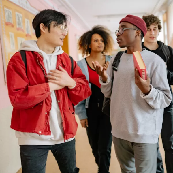 Four students stand in a hallway talking. One holds a book, and all wear casual clothes with backpacks. A bulletin board is visible in the background.