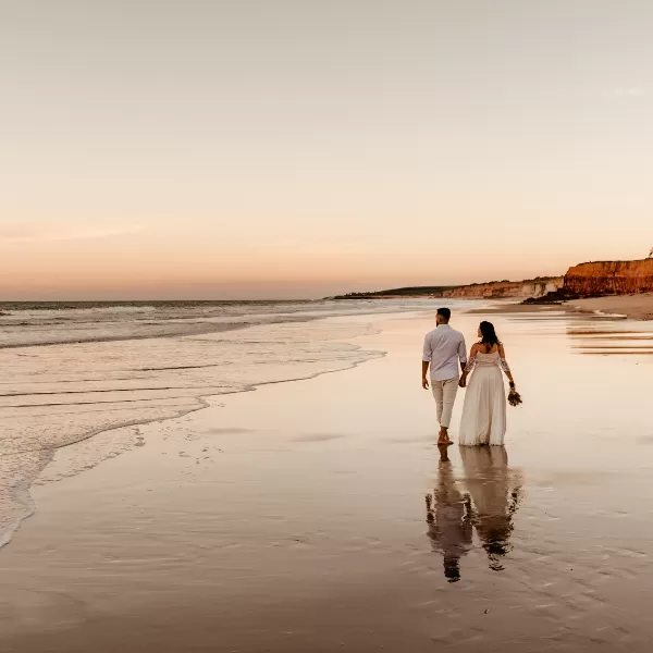 A couple in wedding attire walks barefoot along a beach at sunset, holding hands with gentle waves and cliffs in the background.