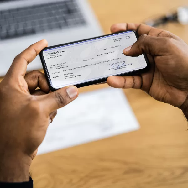 A person holds a smartphone showing a digital check, with a laptop and a paper on a wooden desk in the background.