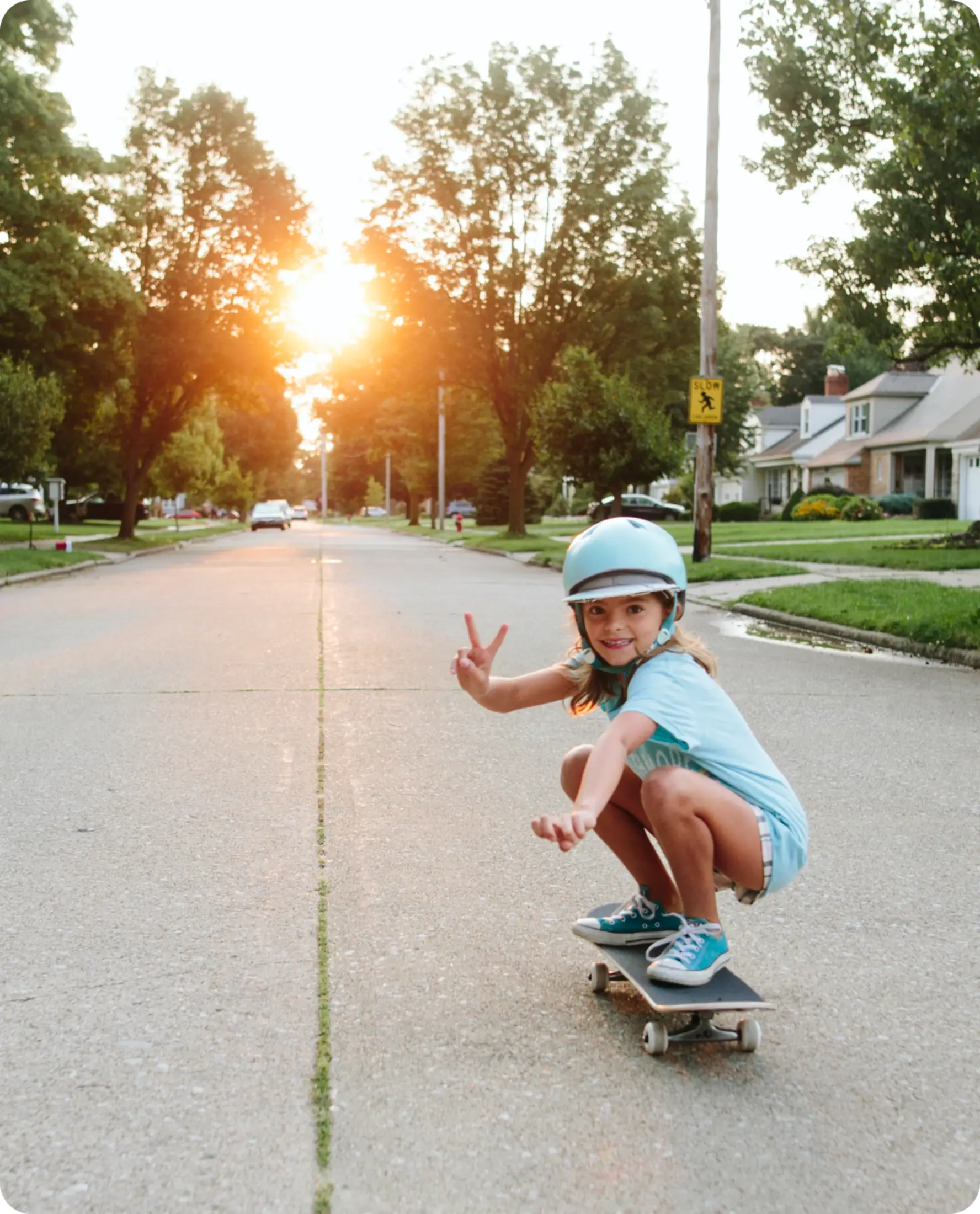 Young child in blue helmet and outfit skateboarding on a suburban street at sunset, smiling and making a peace sign.