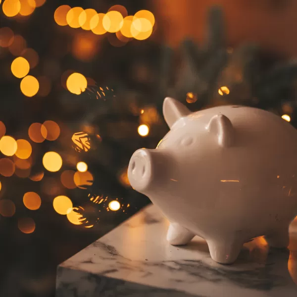 A white ceramic piggy bank on a marble surface with warm, blurred holiday lights in the background.