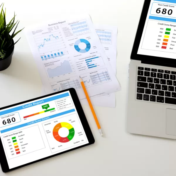 A laptop and tablet display credit score reports of 680, next to printed financial charts and a pencil on a white desk.