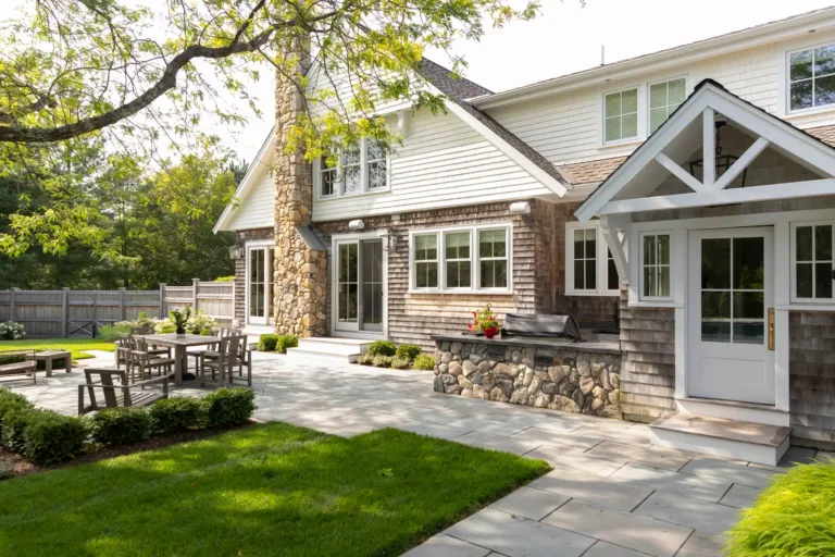 A spacious backyard patio with stone flooring, outdoor dining set, built-in grill, and landscaped lawn beside a two-story house with large windows and stone chimney.
