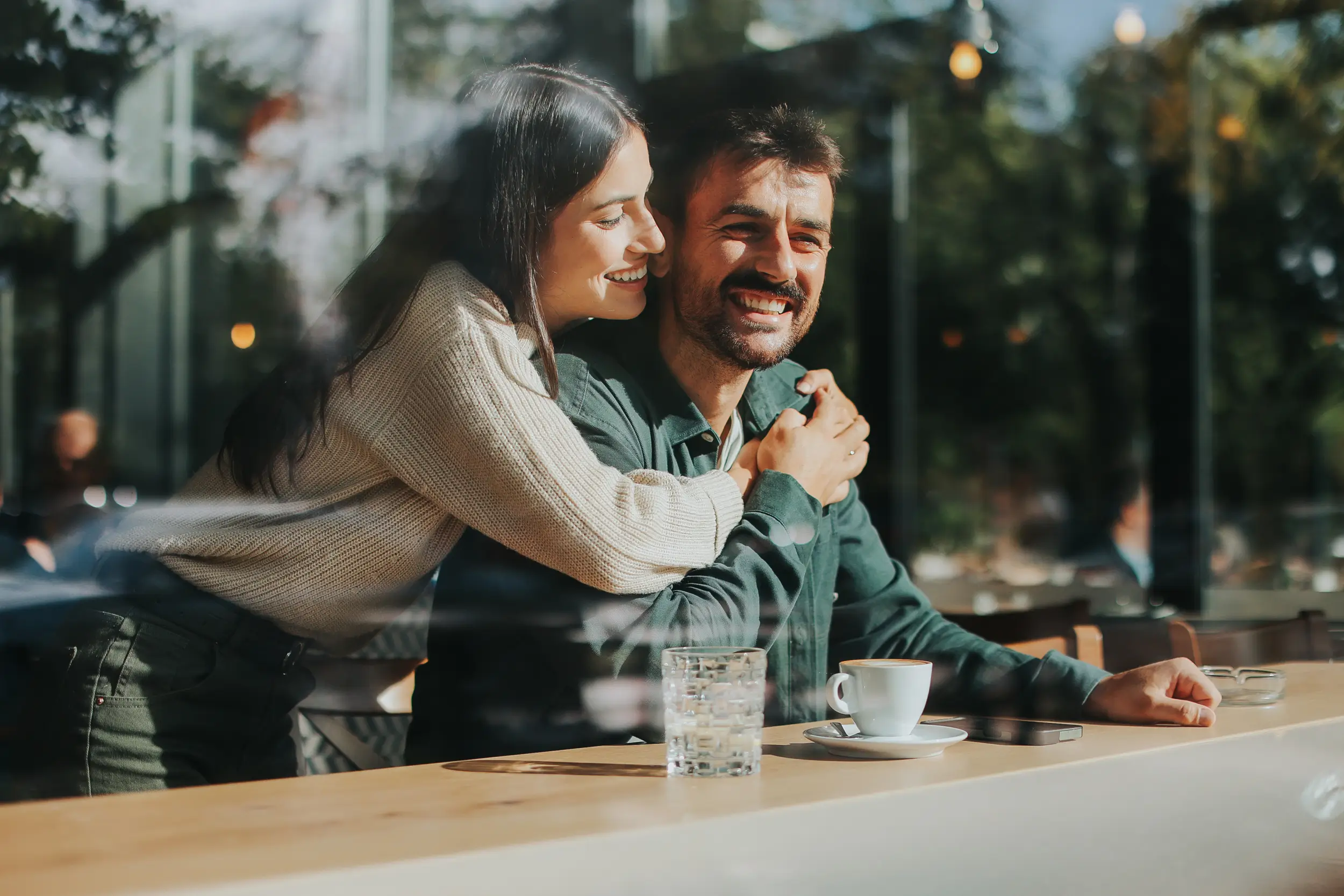 A smiling woman hugs a laughing man from behind as they sit at a café table with a coffee cup and glass of water, seen through a window.