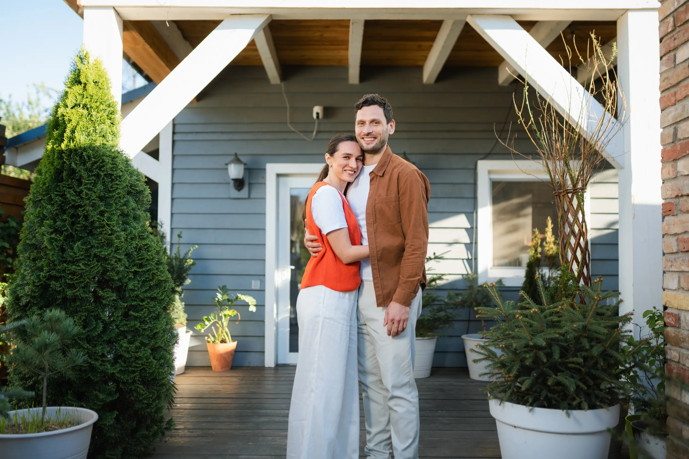 A smiling couple stands hugging on a wooden porch in front of a gray house with white trim and potted plants.