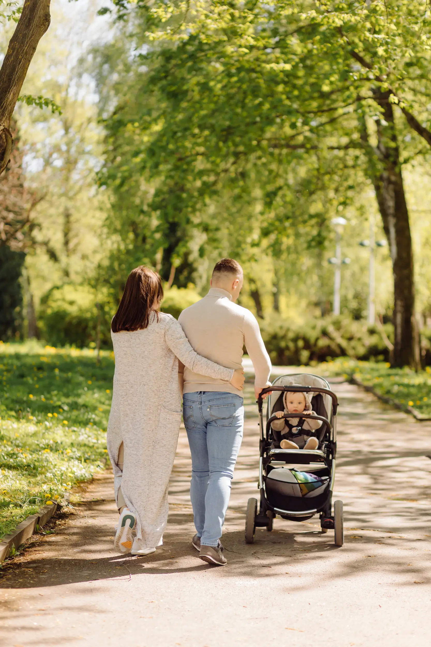A couple walks arm in arm down a sunny park path, pushing a baby in a stroller, surrounded by green trees and grass.