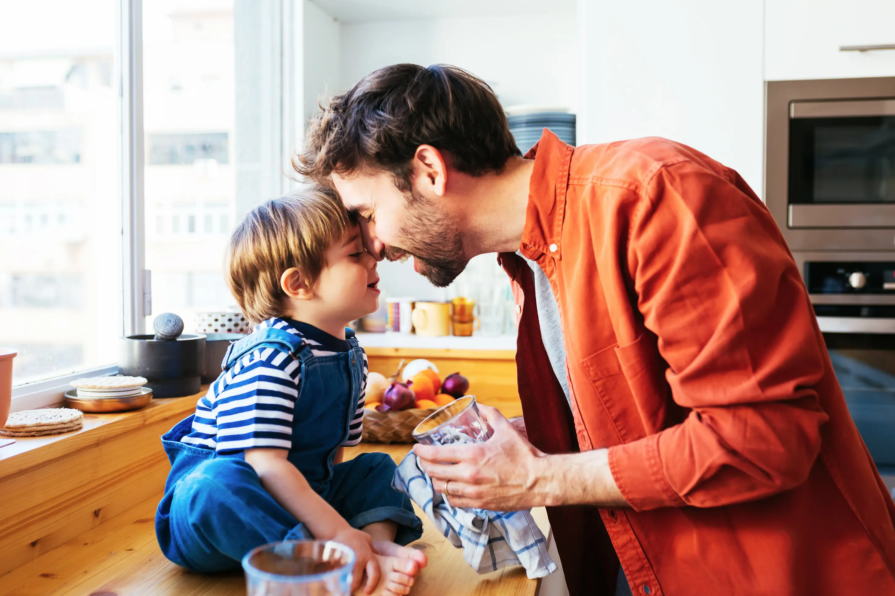 A father gently touches foreheads and smiles with his young child, who is sitting on a kitchen counter in sunlight.