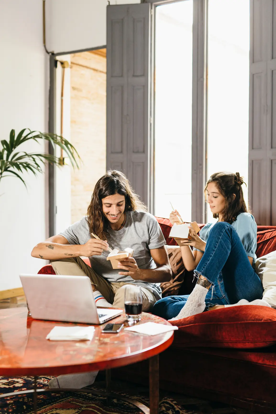 Two people sit on a red couch eating takeout and smiling while watching something on a laptop in a bright living room.