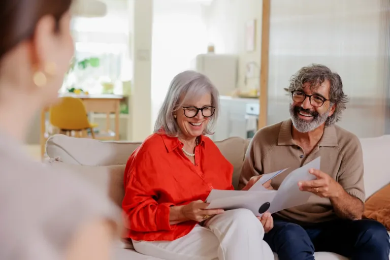 An older couple sits on a couch, smiling and looking at papers, while talking with a person whose back is to the camera in a bright, cozy living room.