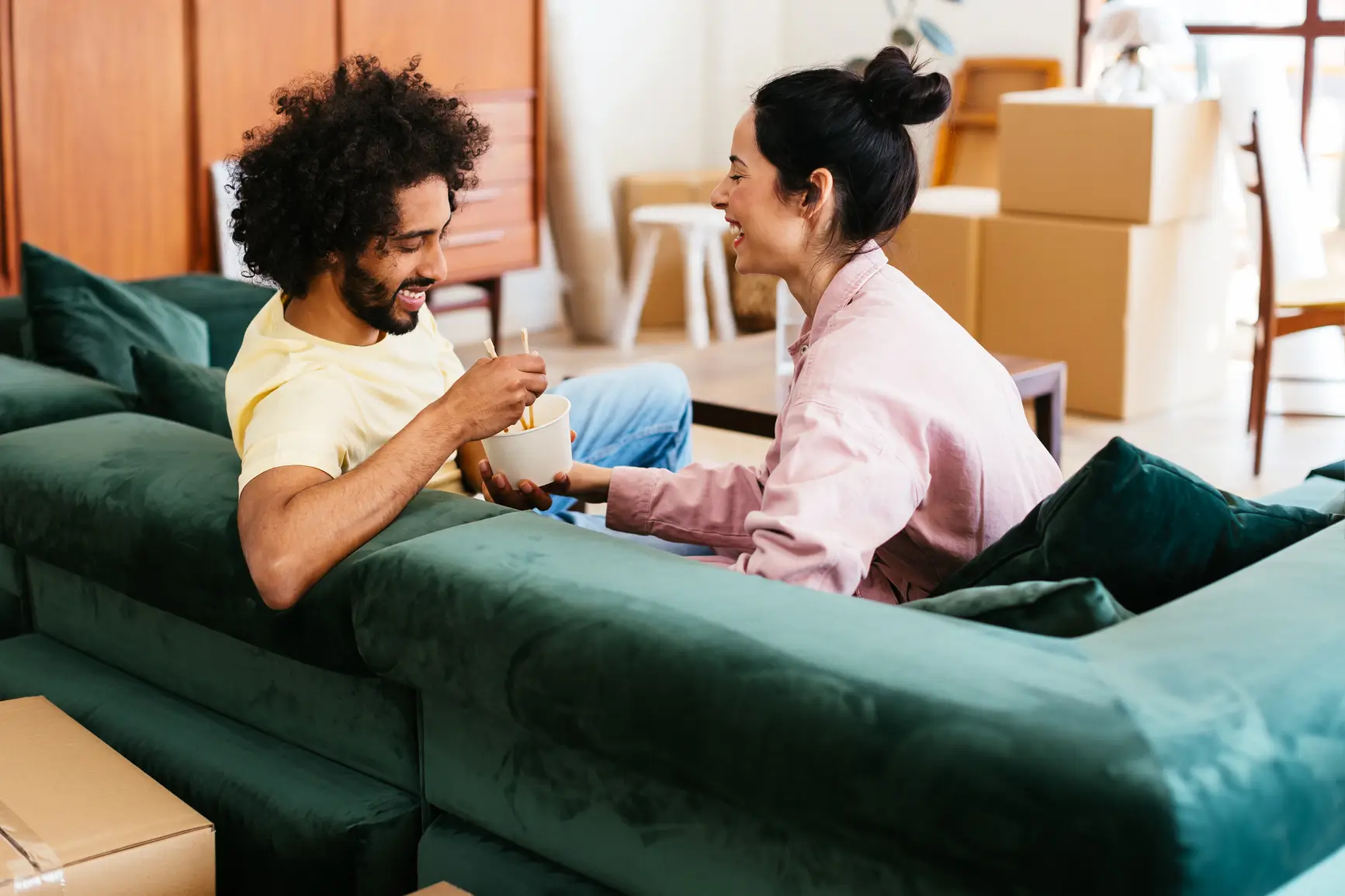 A smiling couple sits on a green sofa, sharing a snack, surrounded by moving boxes in a brightly lit living room.