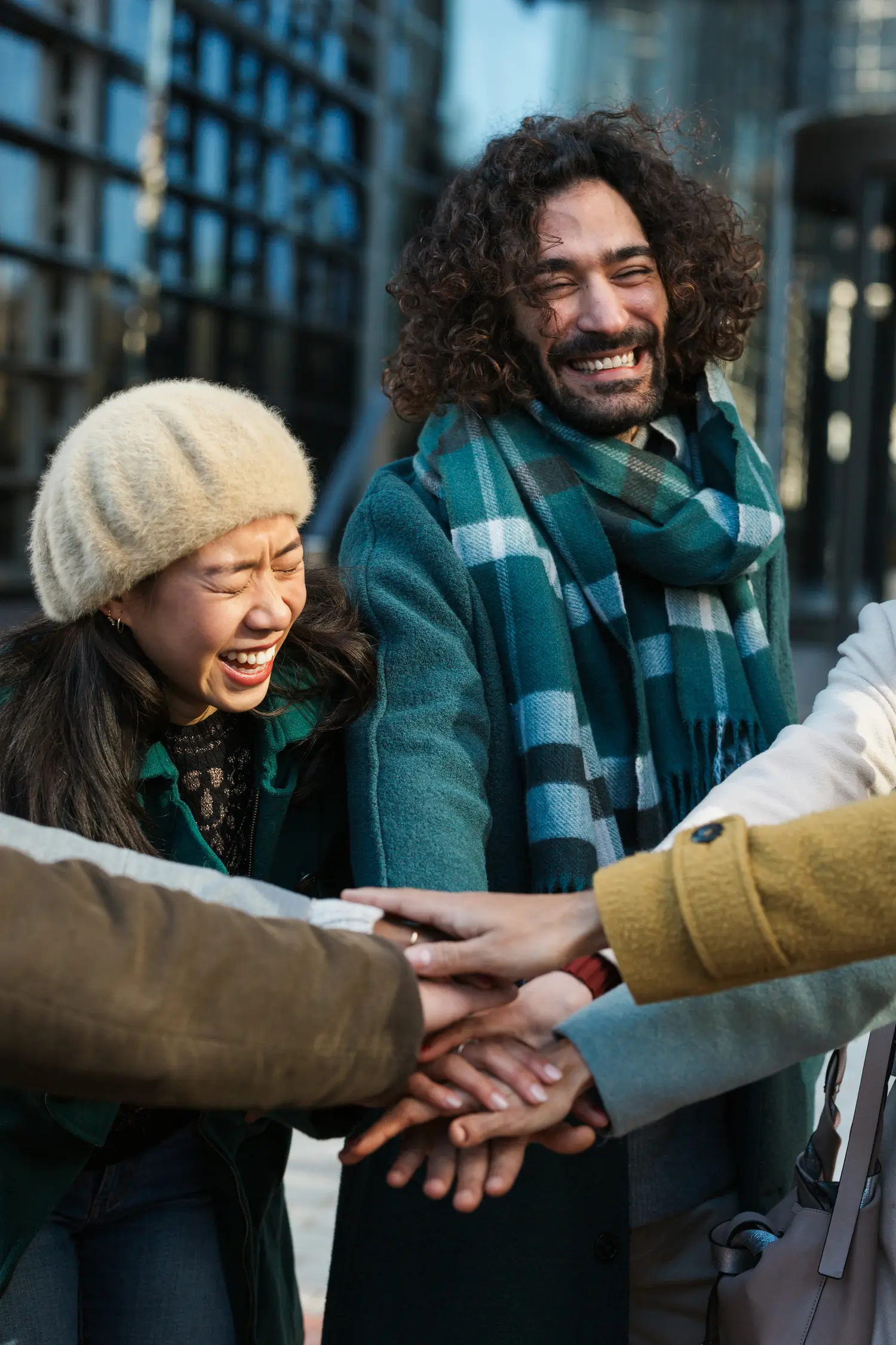 A group of people stand in a circle outdoors, stacking their hands together and smiling, showing unity and friendship.