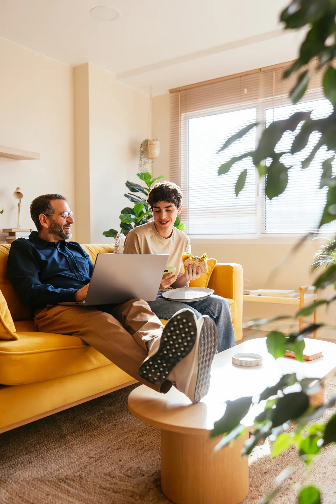 Two people sit on a yellow couch at home; one uses a laptop while the other eats a sandwich and smiles. Sunlight shines through the window, and green plants decorate the room.