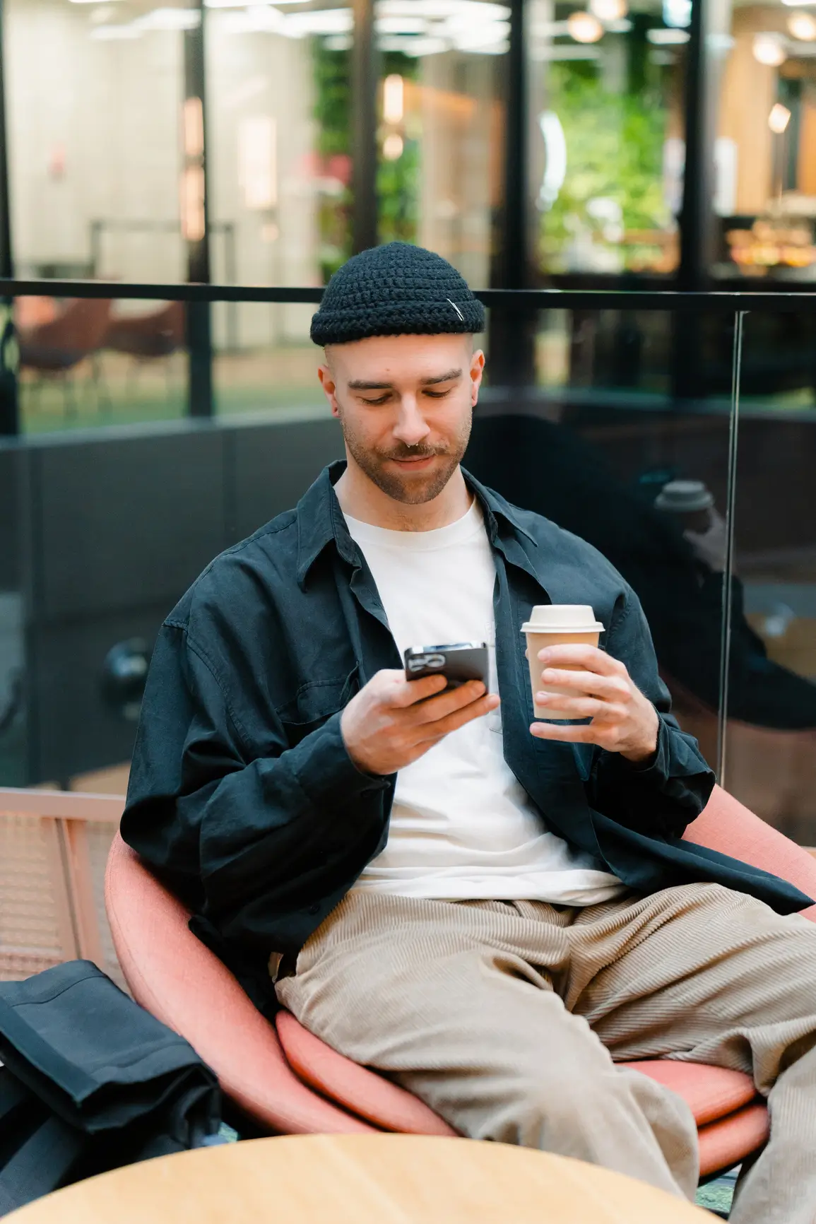 A man wearing a black beanie and jacket sits in a chair, holding a coffee cup in one hand and looking at his phone with a slight smile.