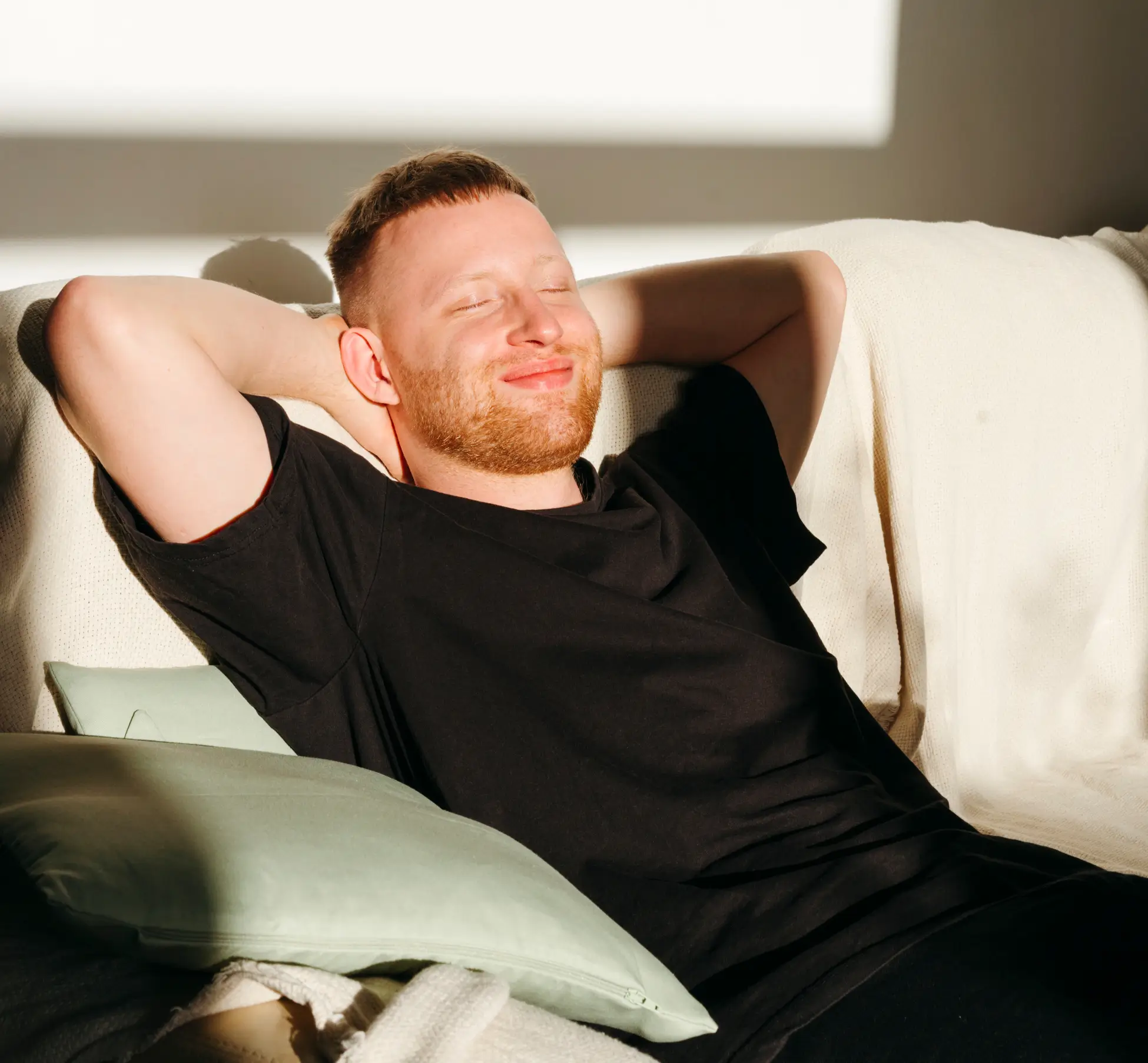 A man with short hair and a beard relaxes on a couch with his arms behind his head, smiling with eyes closed in sunlight. Two cushions are beside him.