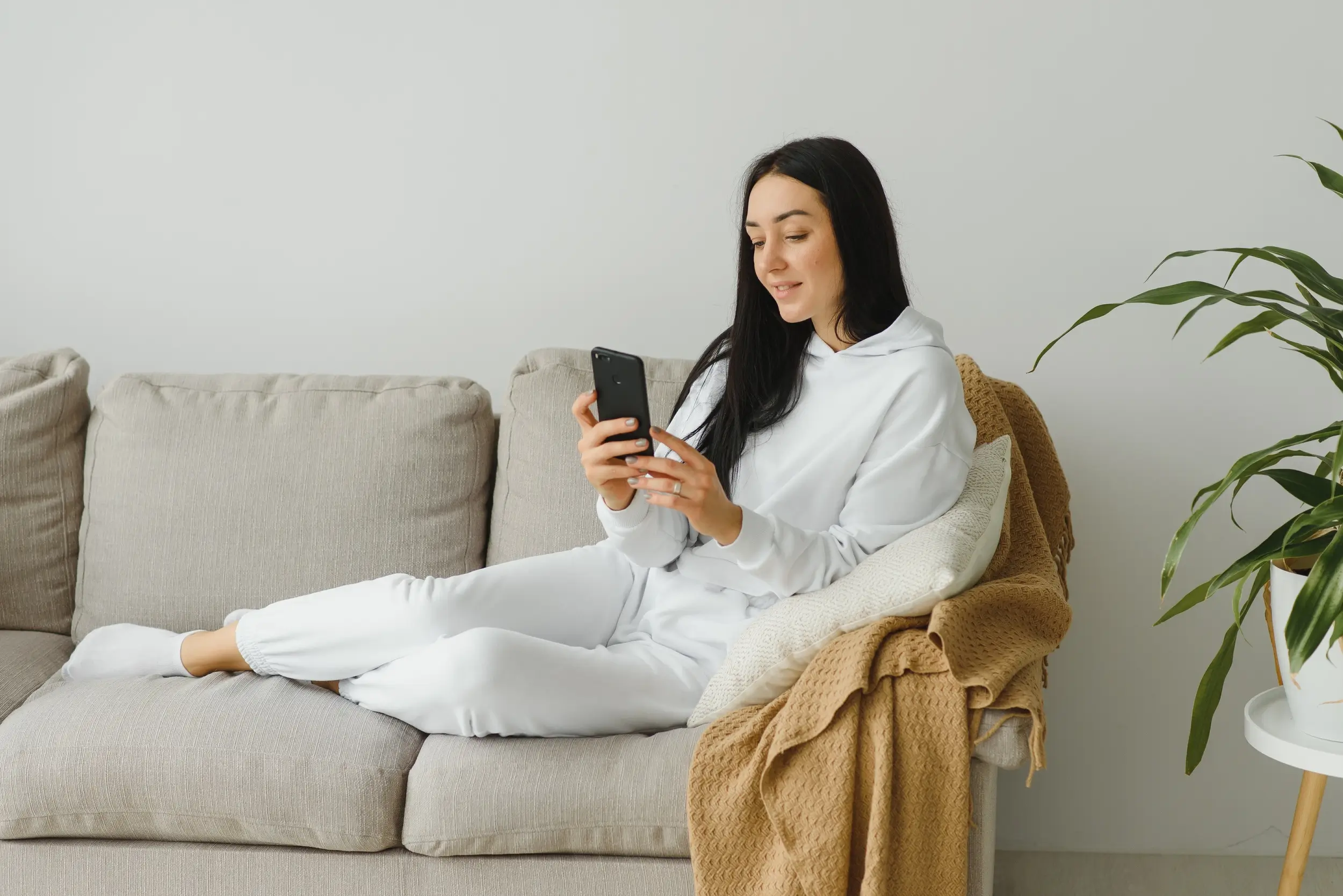 A woman in white loungewear sits on a gray sofa, smiling while looking at her smartphone. A beige blanket and cushion are beside her, with a green plant in the background.