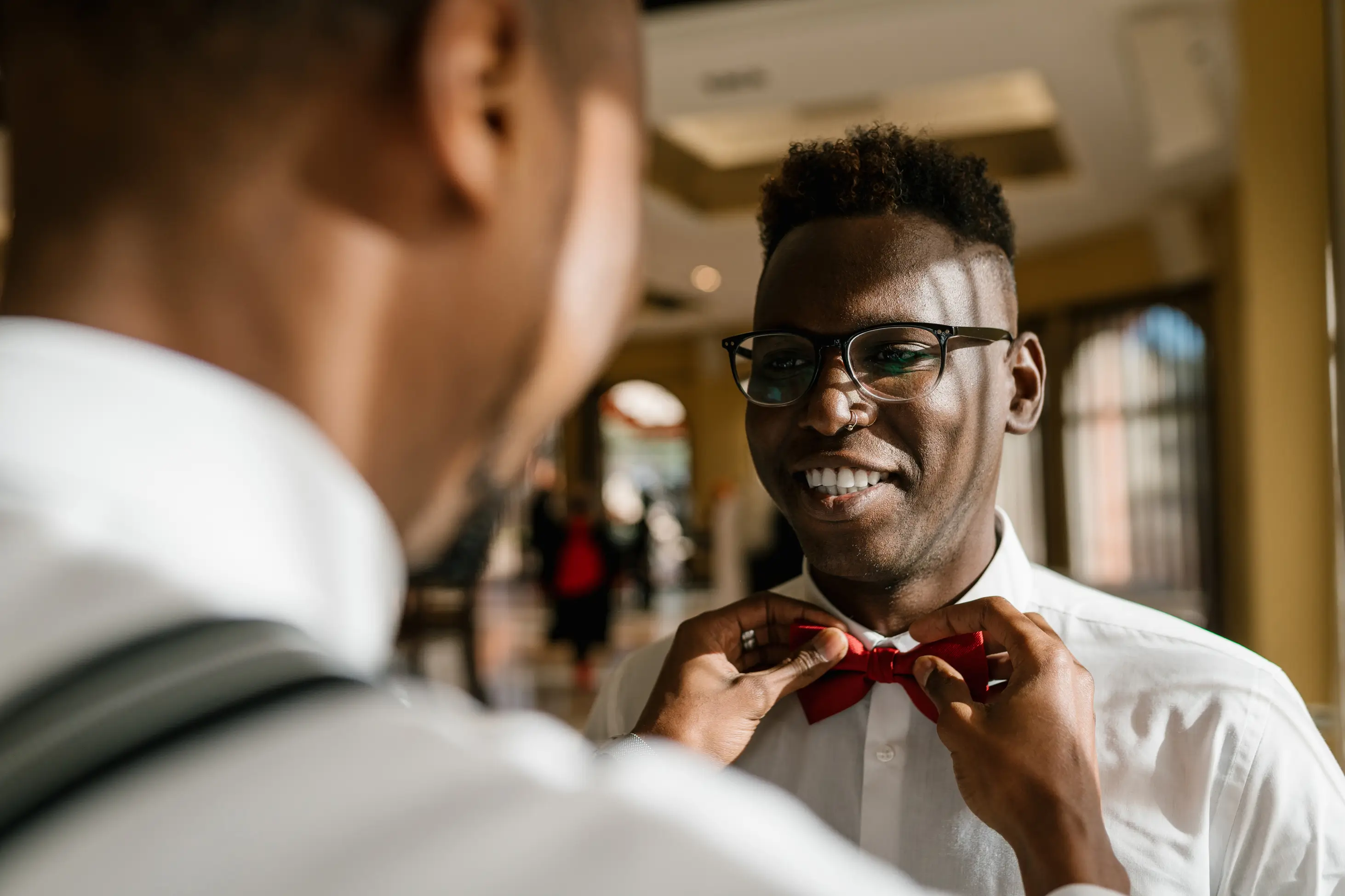 A man smiles as another person helps him adjust a red bow tie; both are dressed formally and standing indoors in a sunlit room.