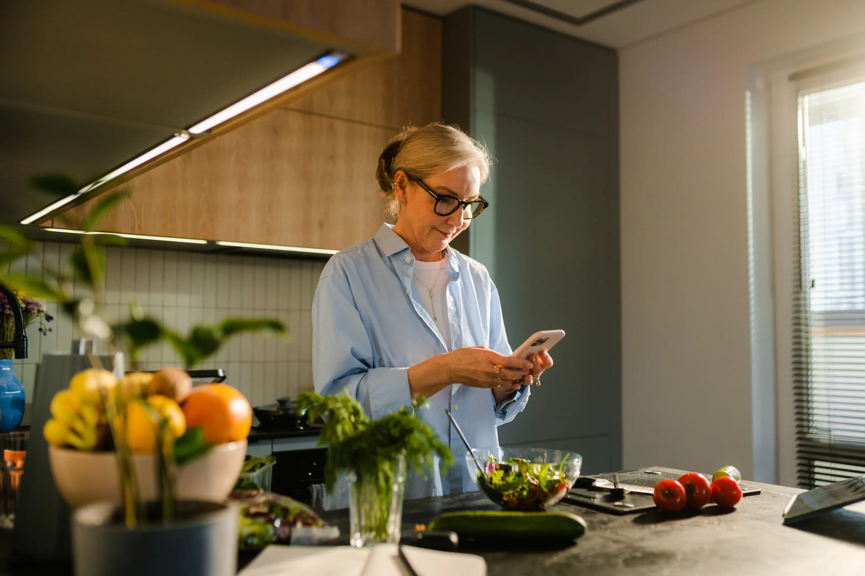 An older woman with glasses stands in a modern kitchen, holding a smartphone and preparing a salad with fresh vegetables on the counter.