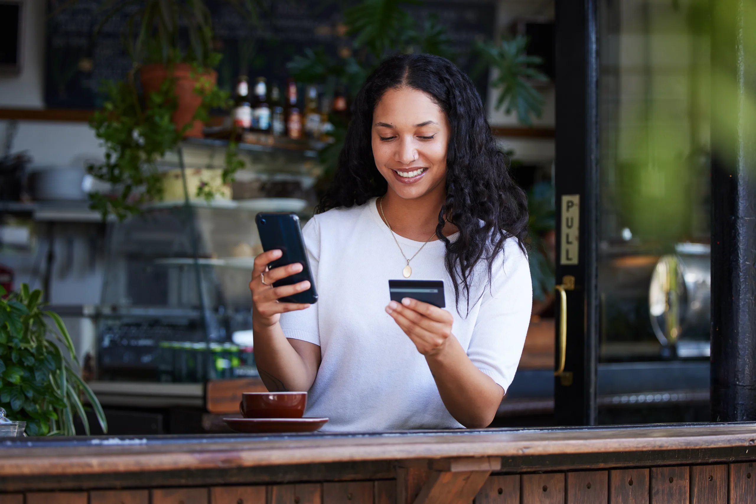 A woman smiles while holding a smartphone and a credit card at a café counter, with a coffee cup in front of her and plants in the background.