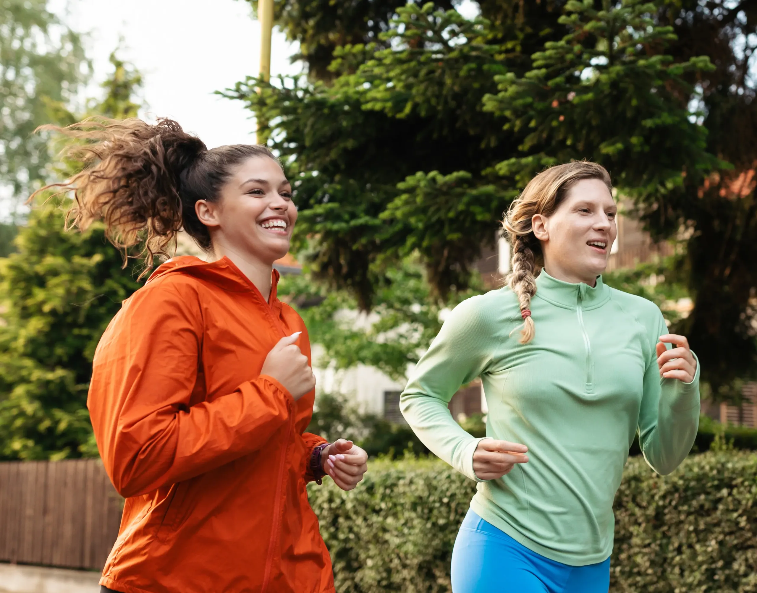 Two women in athletic jackets are jogging outdoors, smiling, surrounded by greenery and trees.