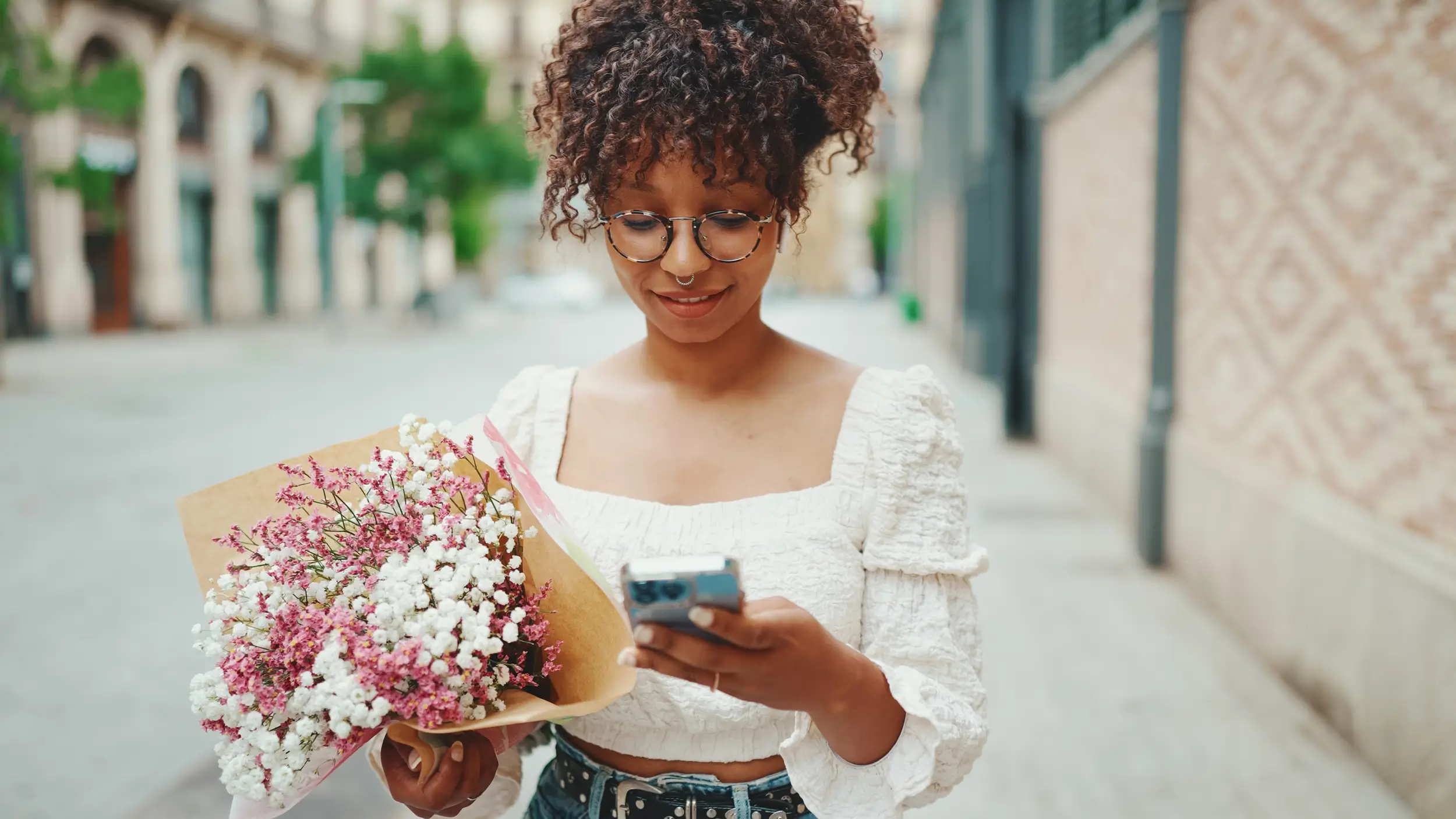 A woman with curly hair and glasses holds a bouquet of flowers and looks at her phone while walking down a city street.