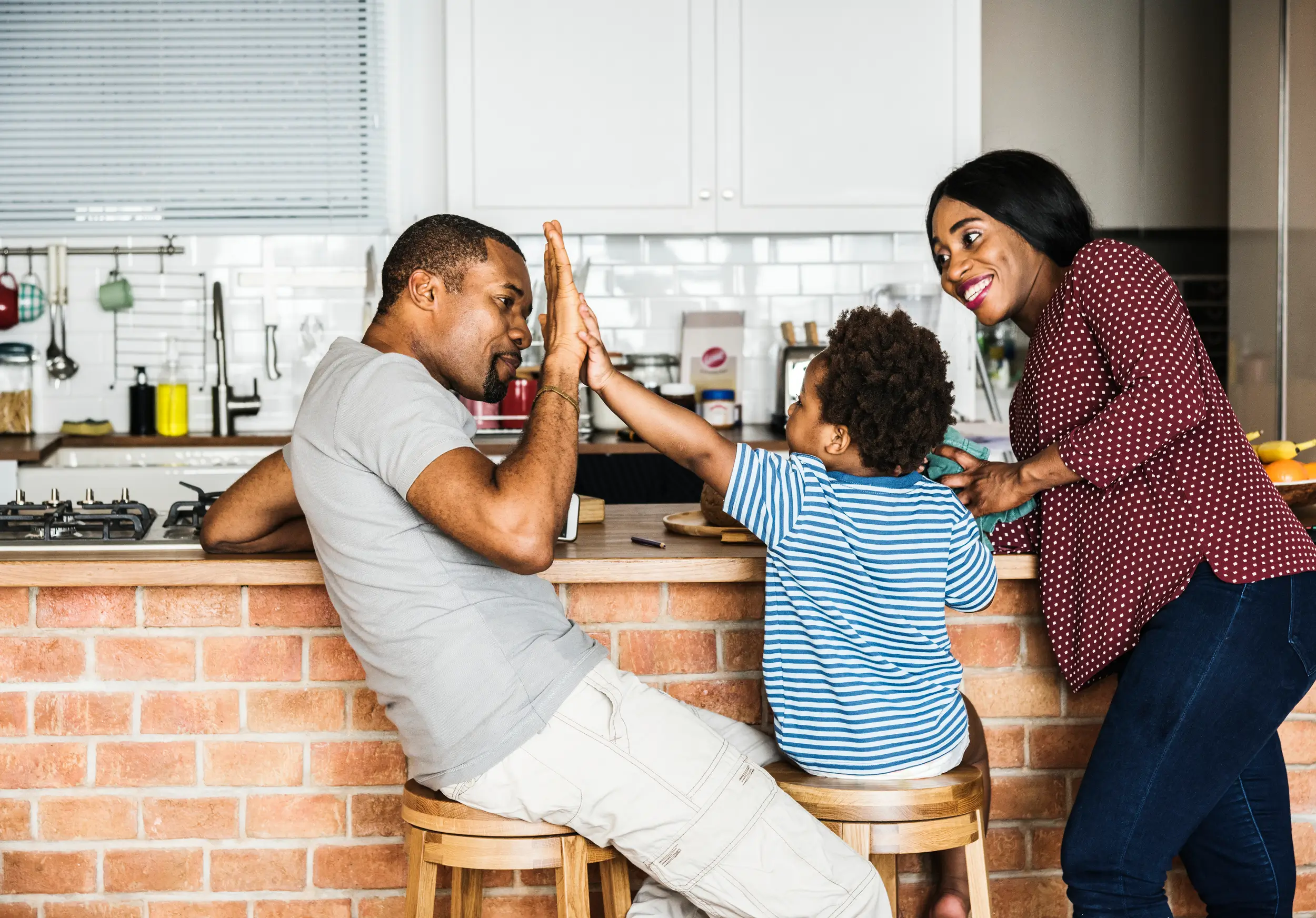 A cheerful family in a kitchen, with a father and young son sharing a high-five while the mother smiles, all sitting at a brick counter.