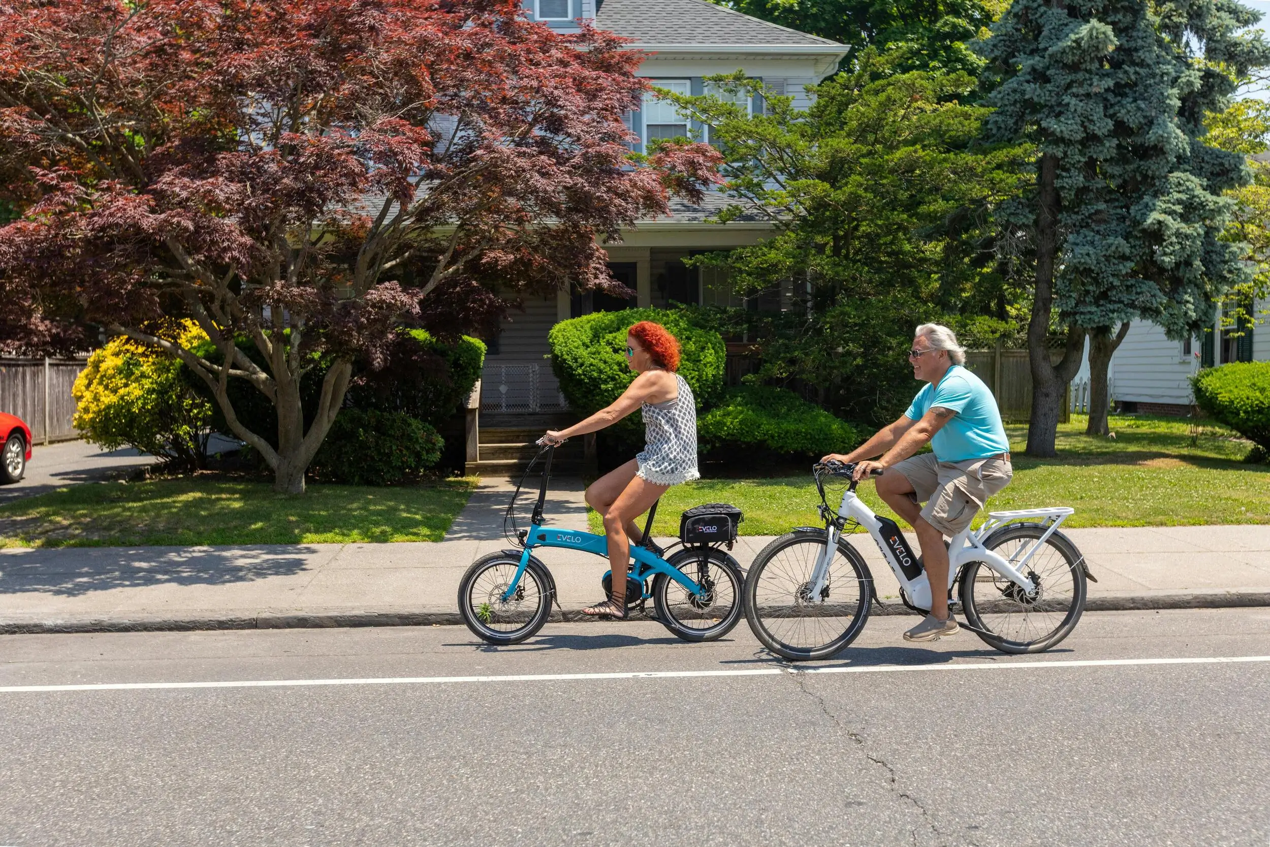 Two people ride electric bikes on a suburban street, passing houses with green lawns and trees on a sunny day.
