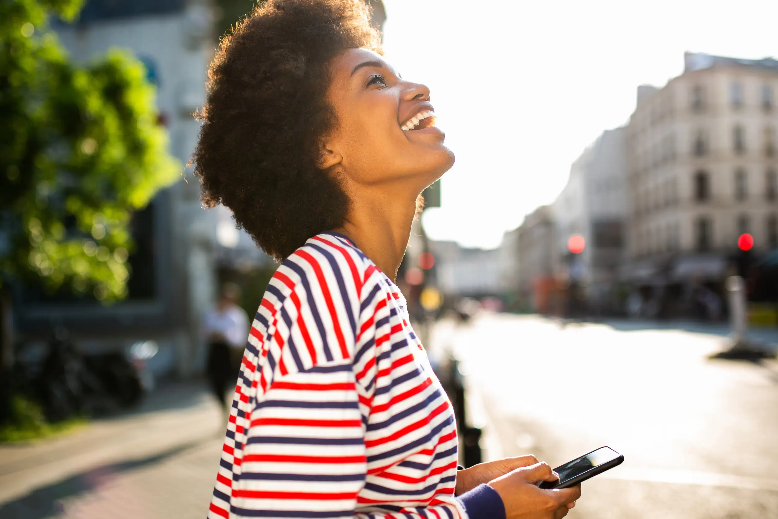 A smiling woman in a striped shirt stands outdoors on a sunny day, holding a smartphone and looking up, with a city street in the background.