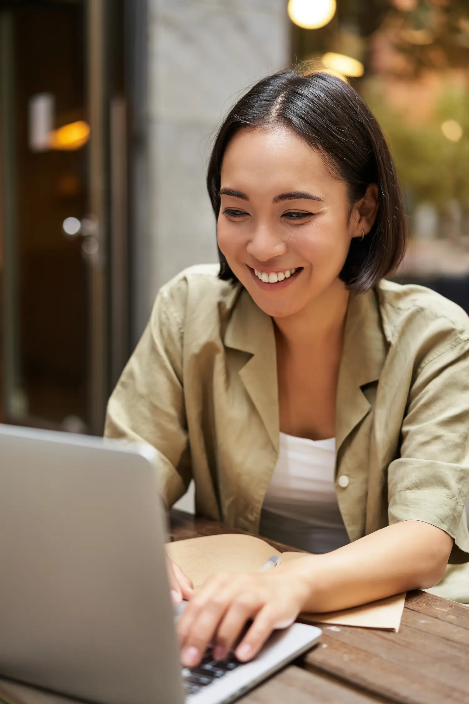 A woman with short dark hair smiles while working on a laptop outdoors at a wooden table.