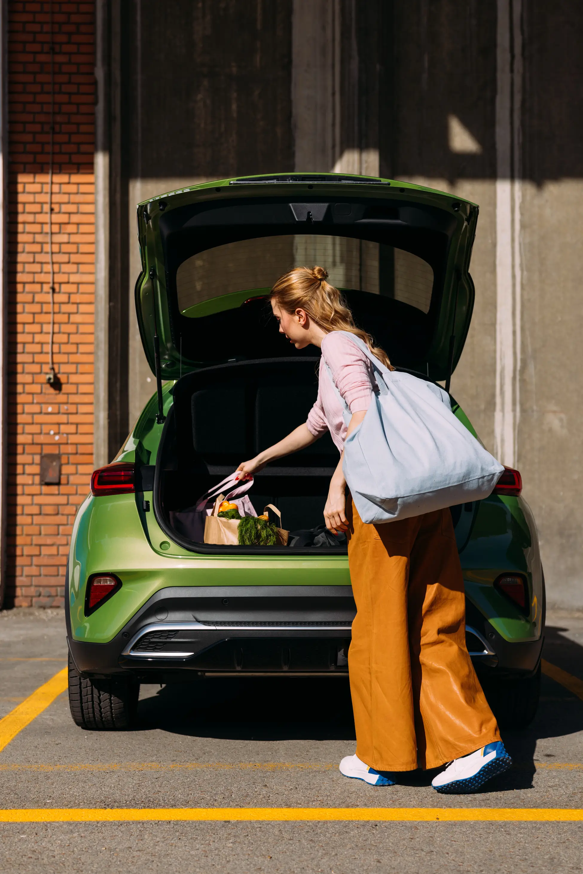 A woman wearing orange pants loads groceries into the trunk of a green car parked in a sunny parking lot.