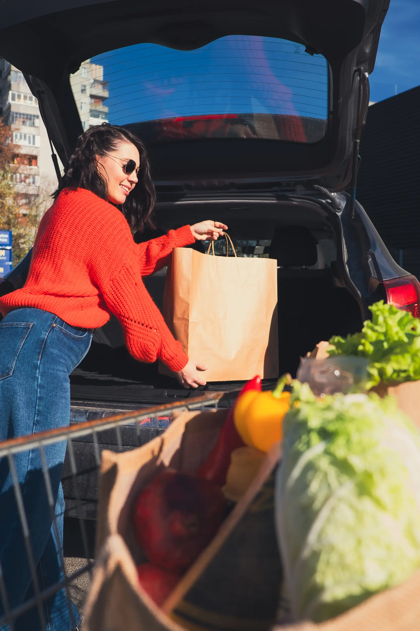 A woman in a red sweater and sunglasses loads a paper grocery bag into the trunk of her car, with fresh produce visible in a shopping cart nearby.