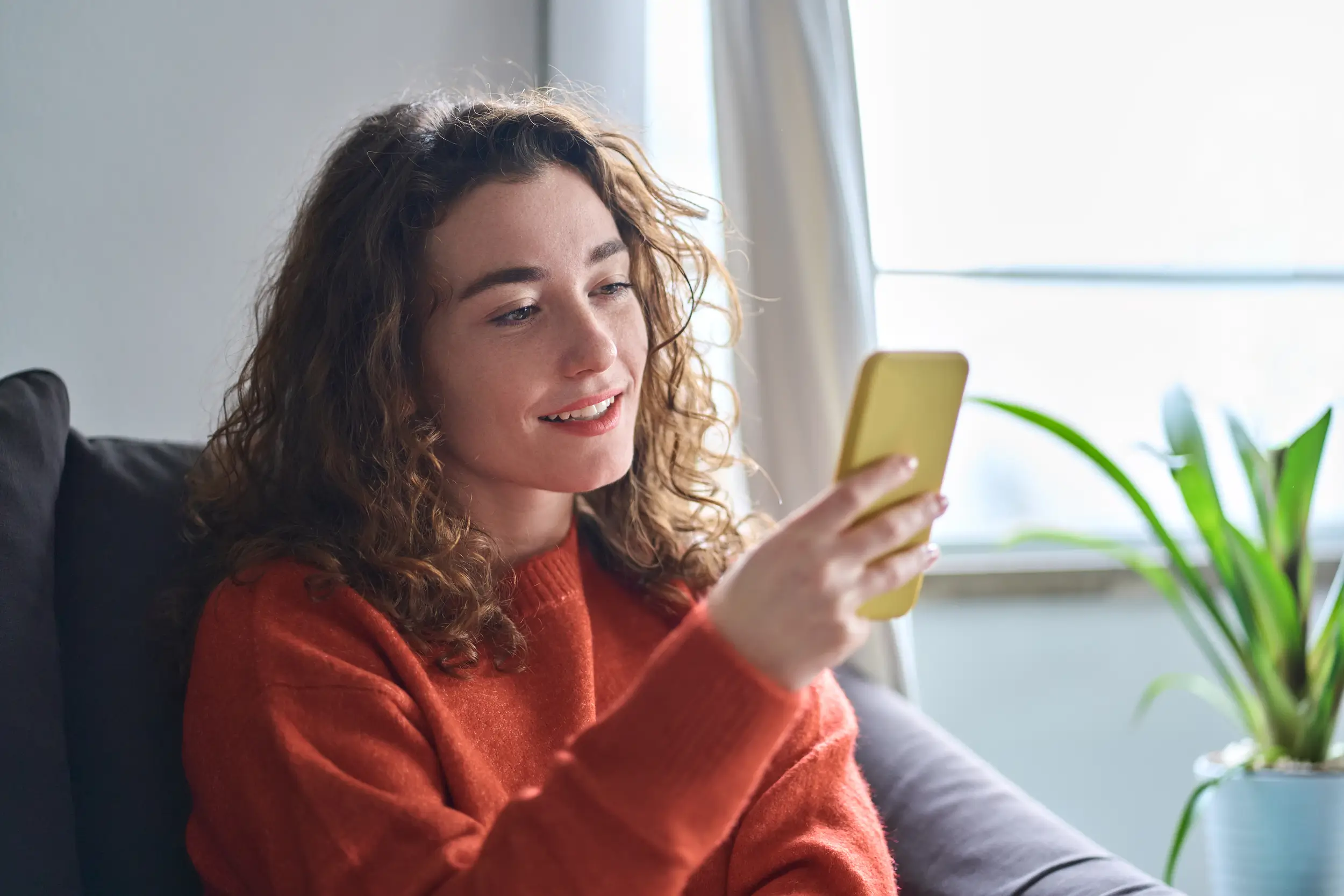 A young woman with curly hair, wearing an orange sweater, sits on a couch smiling while looking at her yellow smartphone. A green plant and window are in the background.