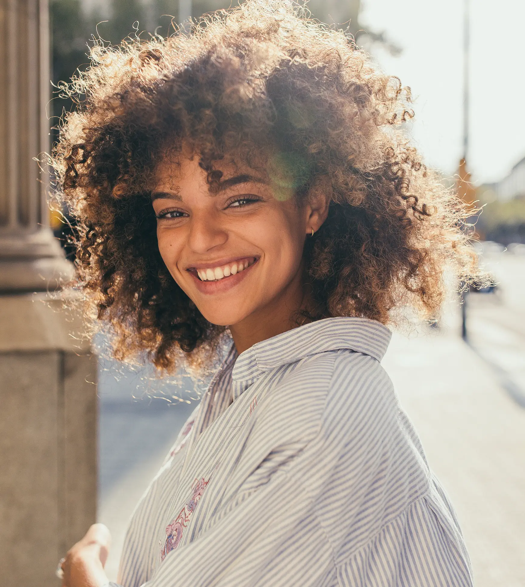 A young woman with curly hair smiles brightly while standing outdoors in sunlight, feeling confident after opening a high yield savings account.