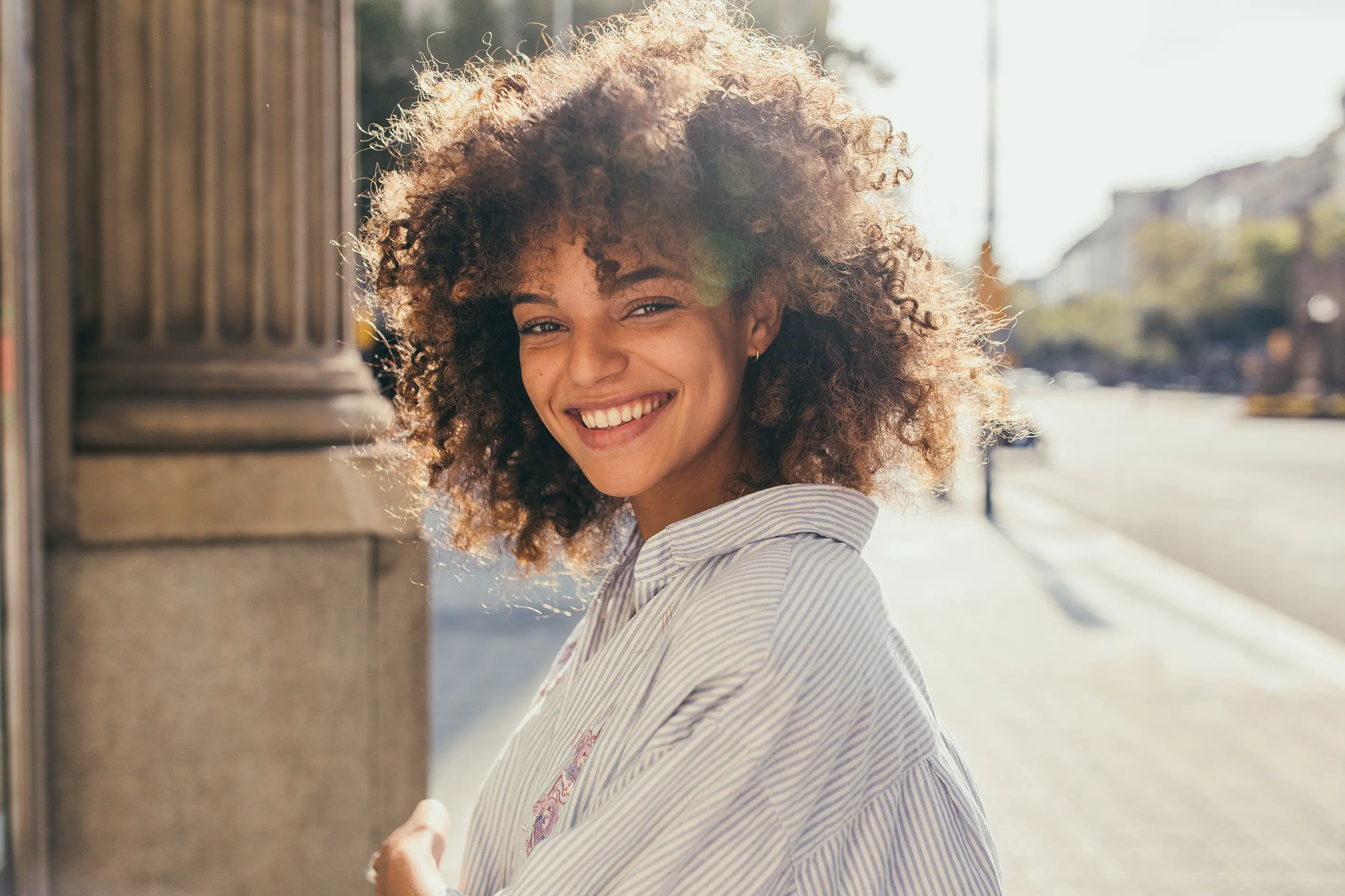 A young woman with curly hair smiles brightly while standing outdoors on a sunny day, wearing a striped shirt.