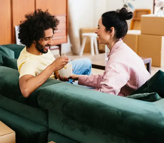 A smiling couple sits on a green couch, sharing a drink and laughing, surrounded by moving boxes in a cozy living room.