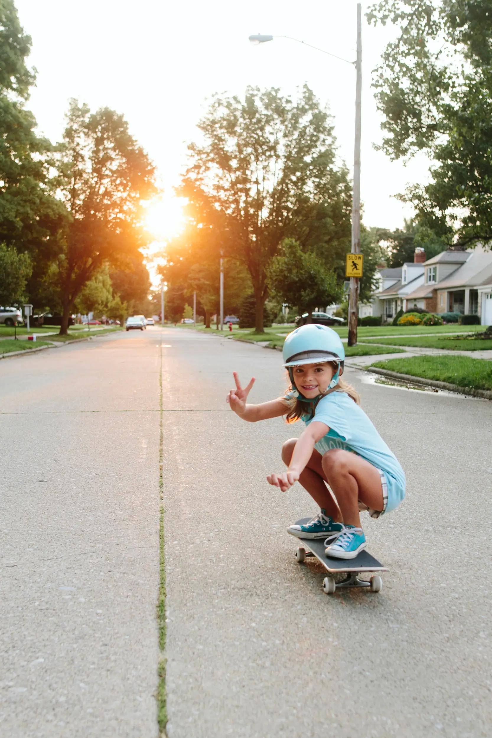 A young girl in a helmet and blue clothes rides a skateboard down a suburban street at sunset, flashing a peace sign—enjoying the freedom, like when you join a credit union.