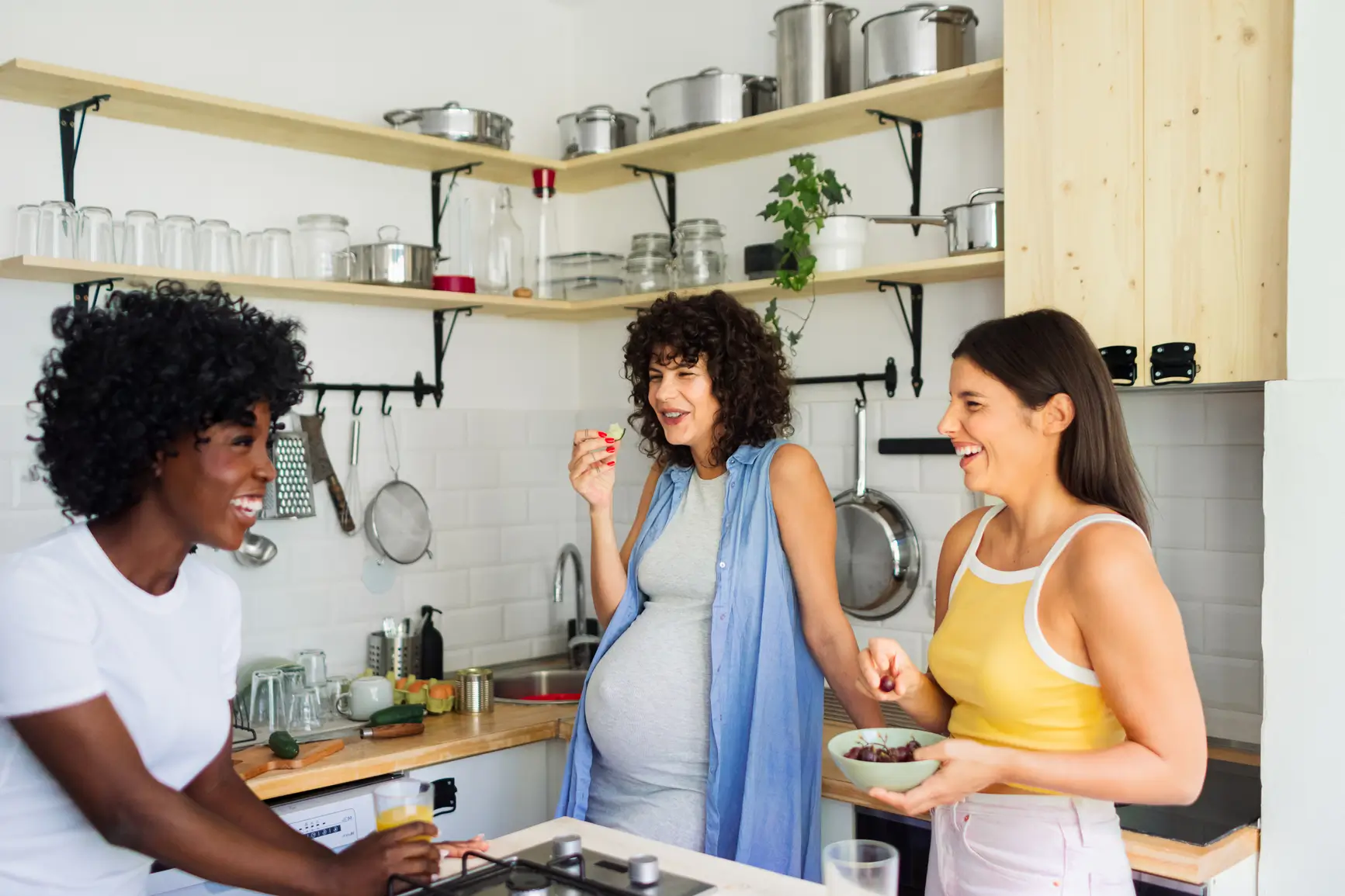 Three women, one visibly pregnant, laugh and talk together in a bright kitchen, enjoying snacks and each others company.