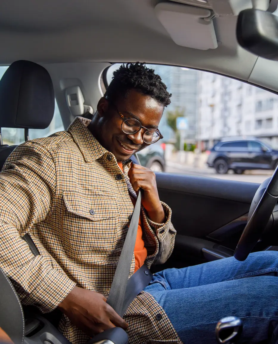 A man sitting in the drivers seat of a car smiles as he fastens his seatbelt. Buildings and parked cars are visible outside the window.