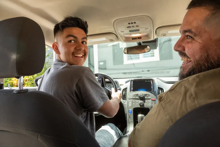 A young man in the driver’s seat and an older man in the passenger seat smile at the camera inside a parked car.