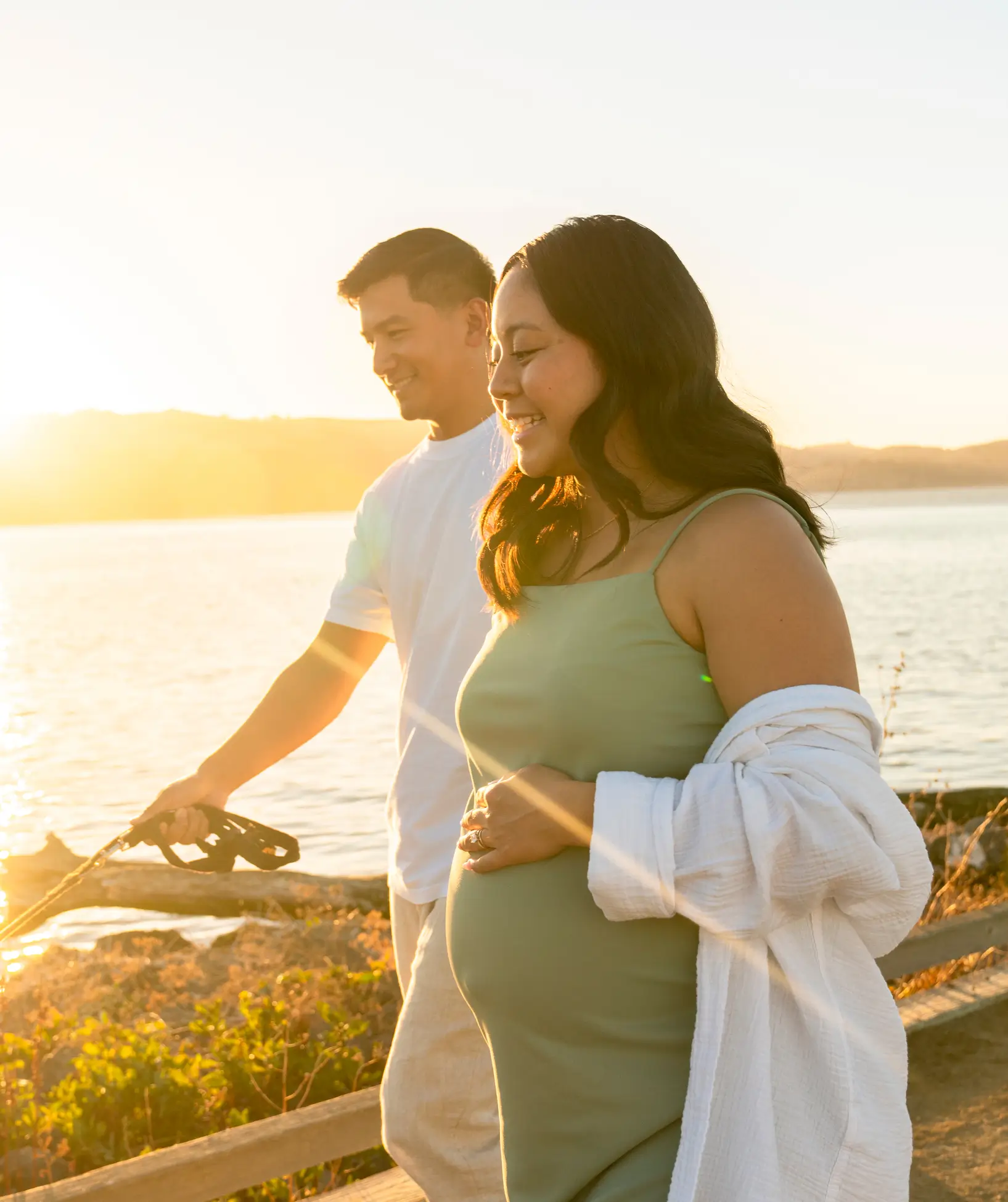 A smiling pregnant woman and a man walk together by the water at sunset, holding hands and enjoying the outdoors.