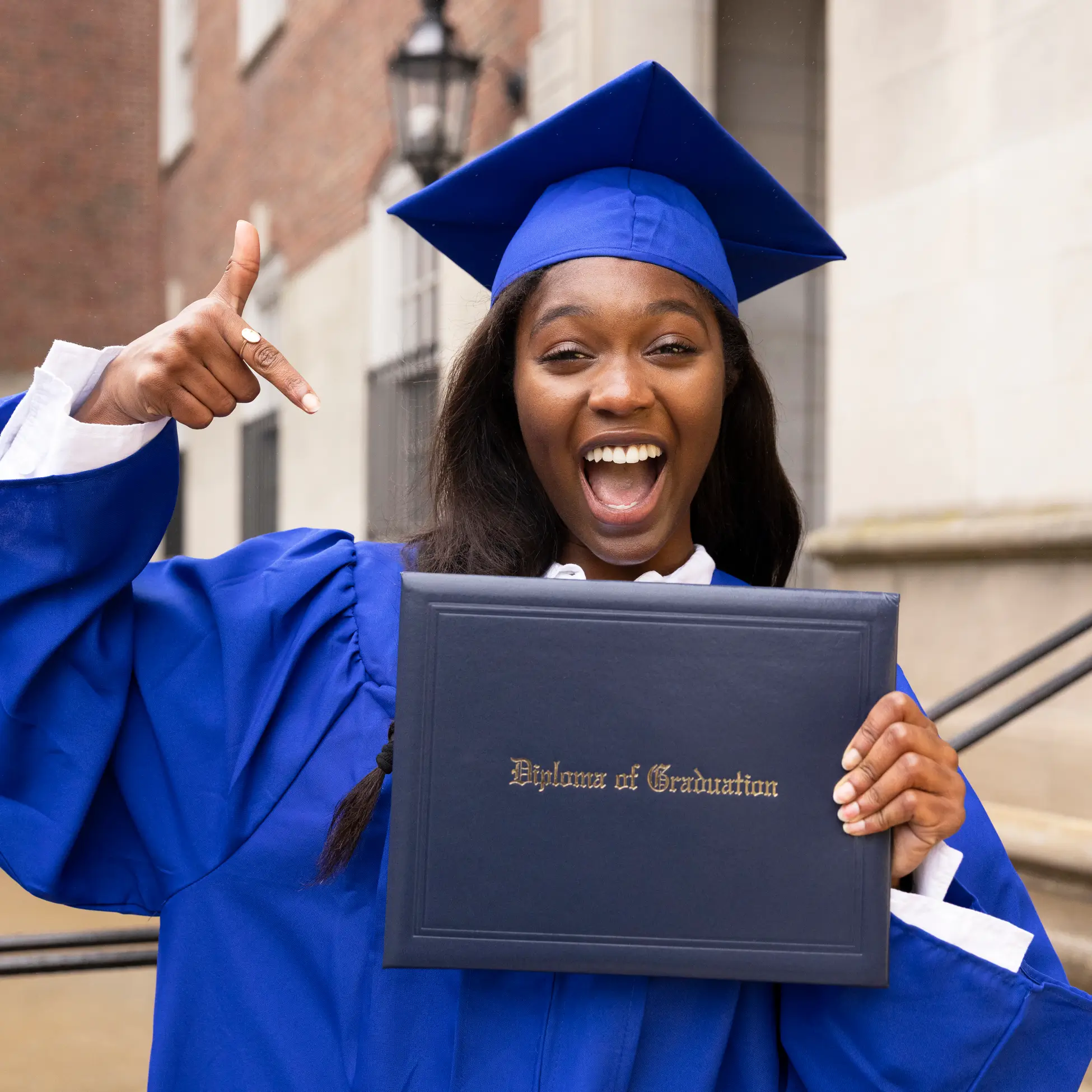 A smiling graduate in a blue cap and gown points excitedly at her diploma while standing outside a building.